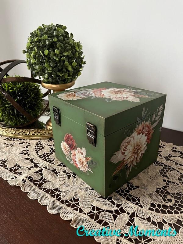 A small, square, green wooden box with metal latches, adorned with floral designs. The box is placed on a lace tablecloth, accompanied by a decorative green ball of faux foliage and a bronze decorative object. Watermark reads "Creative Moments"