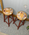 Two wooden stools with round, light brown tops and red legs are adorned with floral wreaths. The stools are placed on a patterned rug in a cozy indoor setting with a plant and a logo in the background.