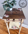 A small square wooden side table with a dark brown top featuring floral designs in white, pink, and red. The table has a single drawer with matching floral artwork and a lower shelf. A potted plant and glasses rest on the table. "AJs Vintage Designs" logo is visible.