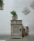 Grayish beige painted nightstand with texture and distressing on the front. The Gilded Damask Transfer has been applied on the front of the piece in the insert of the cabinet door. Staged with a concrete planter containing a faux plant on top. Two large candlesticks on the floor next to the piece with two white candles on top.