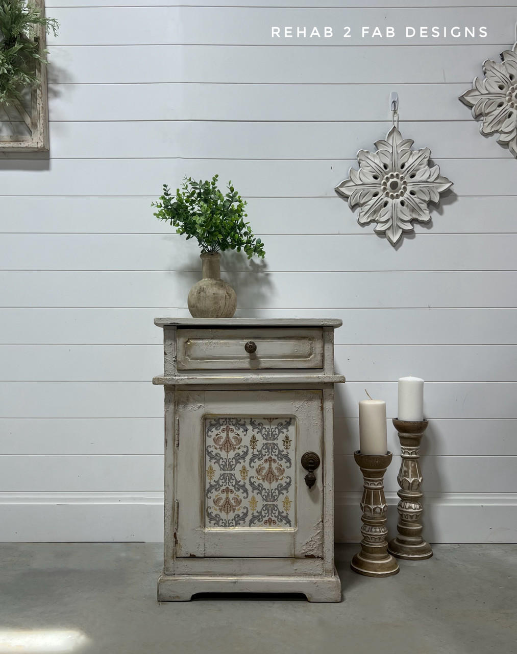 Grayish beige painted nightstand with texture and distressing on the front. The Gilded Damask Transfer has been applied on the front of the piece in the insert of the cabinet door. Staged with a concrete planter containing a faux plant on top. Two large candlesticks on the floor next to the piece with two white candles on top.