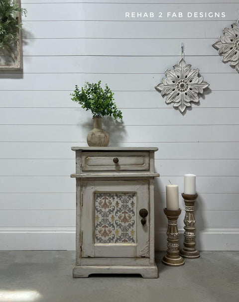 Grayish beige painted nightstand with texture and distressing on the front. The Gilded Damask Transfer has been applied on the front of the piece in the insert of the cabinet door. Staged with a concrete planter containing a faux plant on top. Two large candlesticks on the floor next to the piece with two white candles on top.