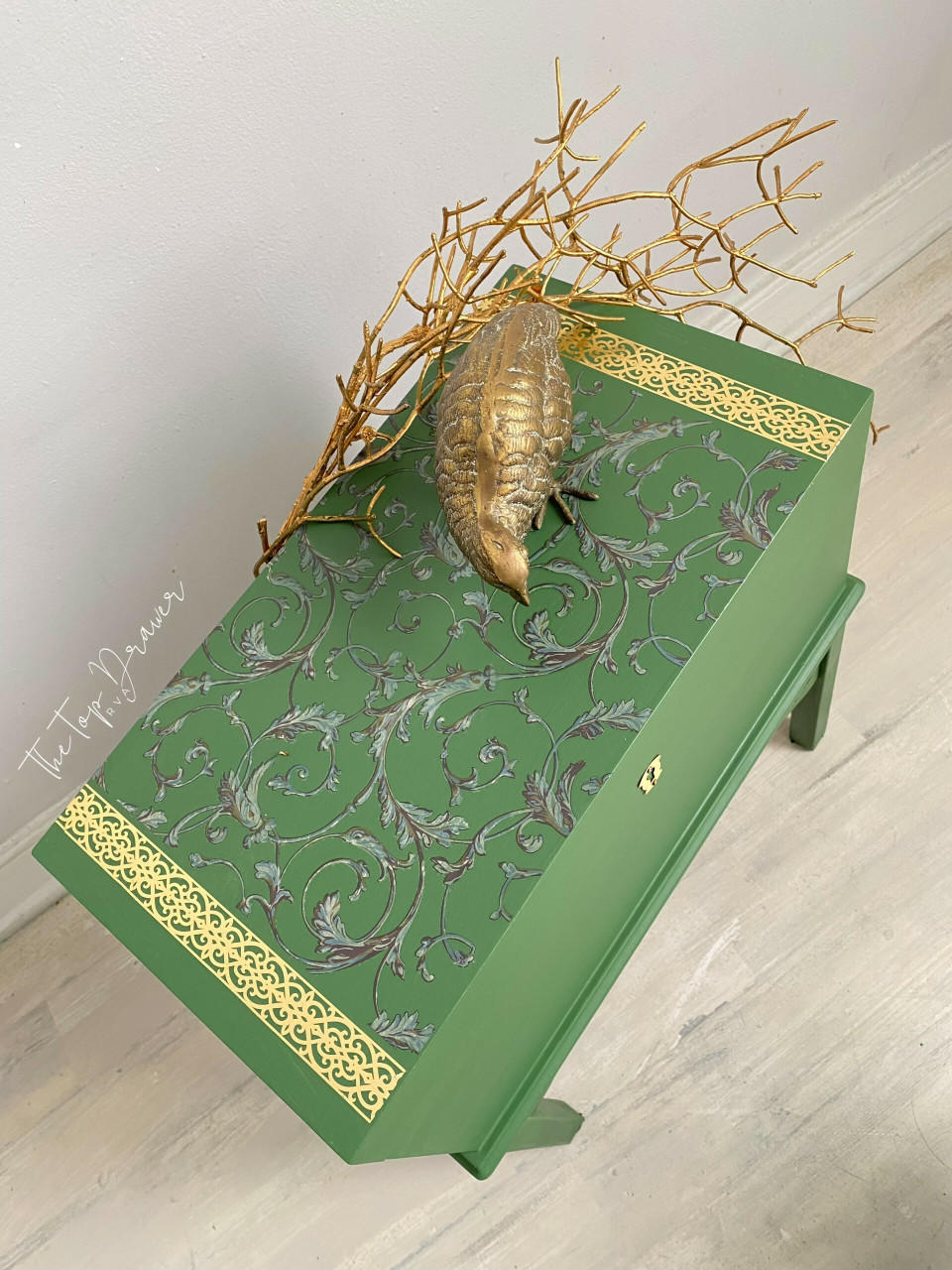 Green painted chest with a gold keyhole on the front. The Baroque Swirls Transfer has been applied on the top with the gold borders on each side. Staged with gold sticks and a gold bird statue on top.