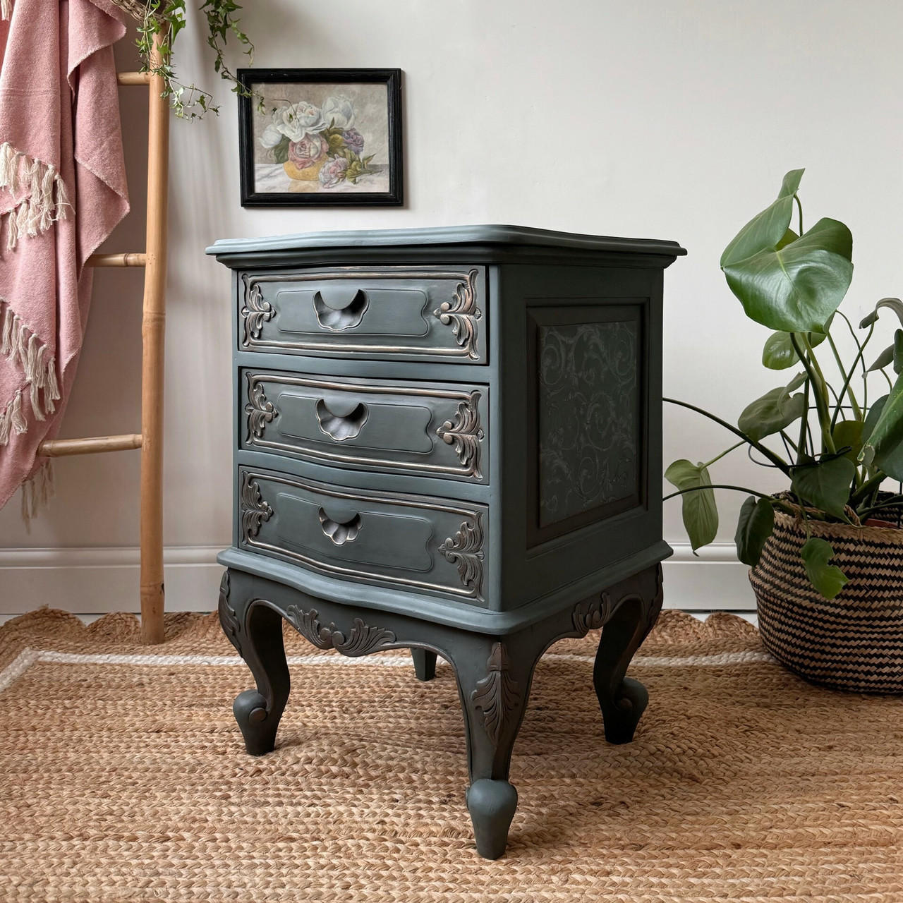 Dark bluish black painted nightstand with the Baroque Swirls Transfer applied on the side of the piece in the insert. Staged on a brown woven rug next to a large monstera plant in a woven potholder with a wooden blanket ladder in the background.
