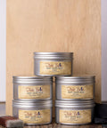 Five metal tins of Dixie Belle Best Dang Wax are stacked in a pyramid against a wooden background. The labels display different colors: Grunge Gray, Brown, Clear, Black and White. A paintbrush rests beside the tins.