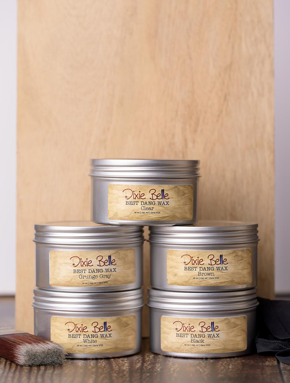 Five metal tins of Dixie Belle Best Dang Wax are stacked in a pyramid against a wooden background. The labels display different colors: Grunge Gray, Brown, Clear, Black and White. A paintbrush rests beside the tins.