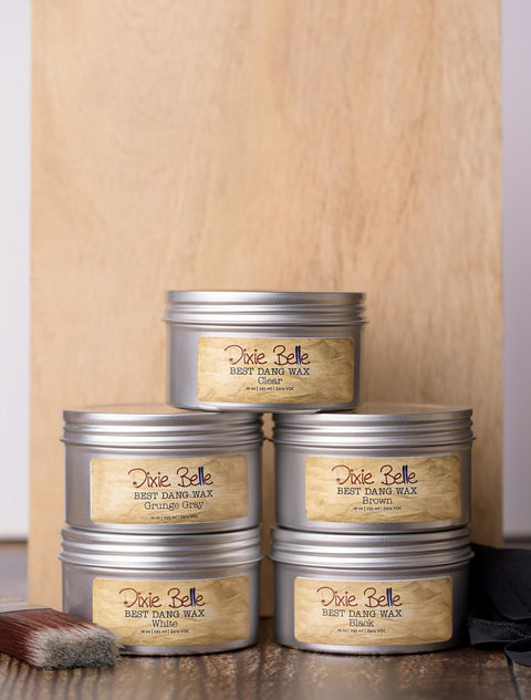 Five metal tins of Dixie Belle Best Dang Wax are stacked in a pyramid against a wooden background. The labels display different colors: Grunge Gray, Brown, Clear, Black and White. A paintbrush rests beside the tins.