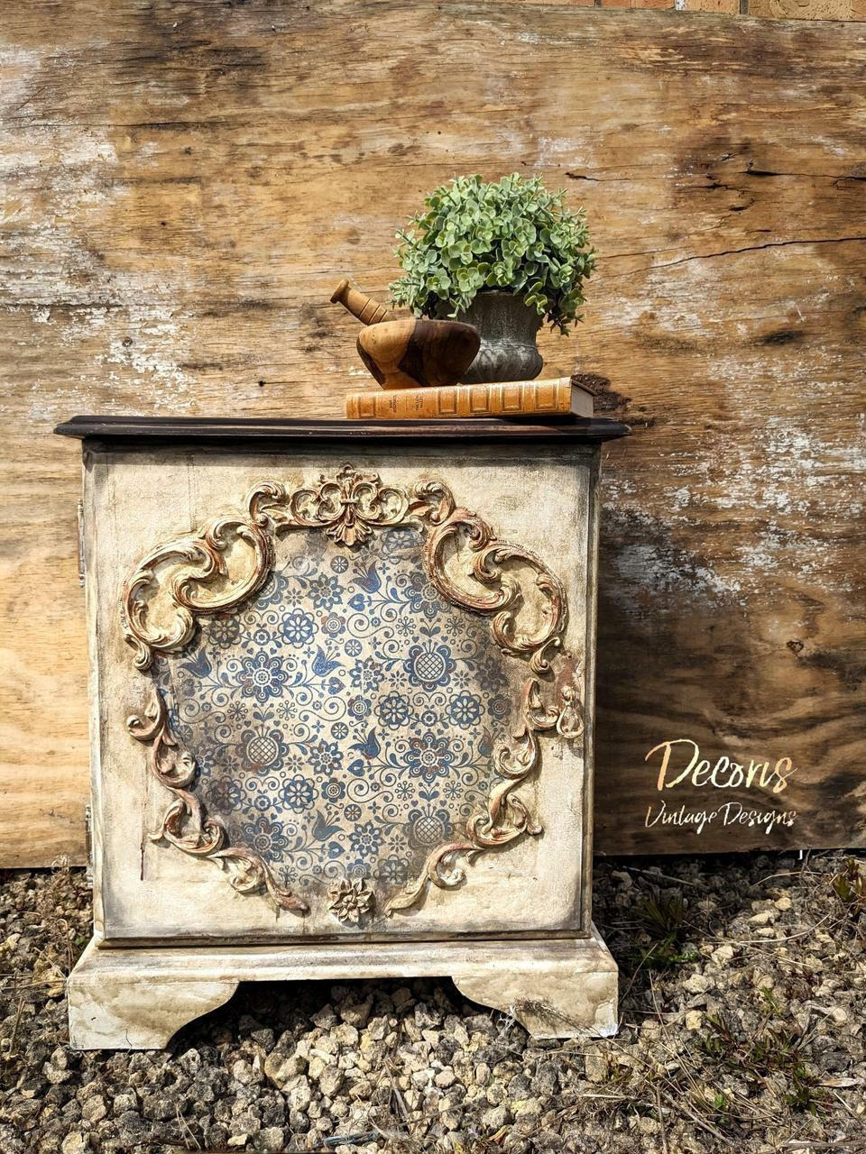 Belles and Whistles Blue Glass Ornate - Rice Decoupage Paper is a simple way to visually enhance any surface of your project! Vintage off-white distressed Side table with Blue glass Ornate rice Decoupage paper used in the center of table inside woodwork. With brown wax to look antique. Staged on top of gravel and Infront of a wood wall with a metal planter and a mortar on top a brown book on the top right corner of table.