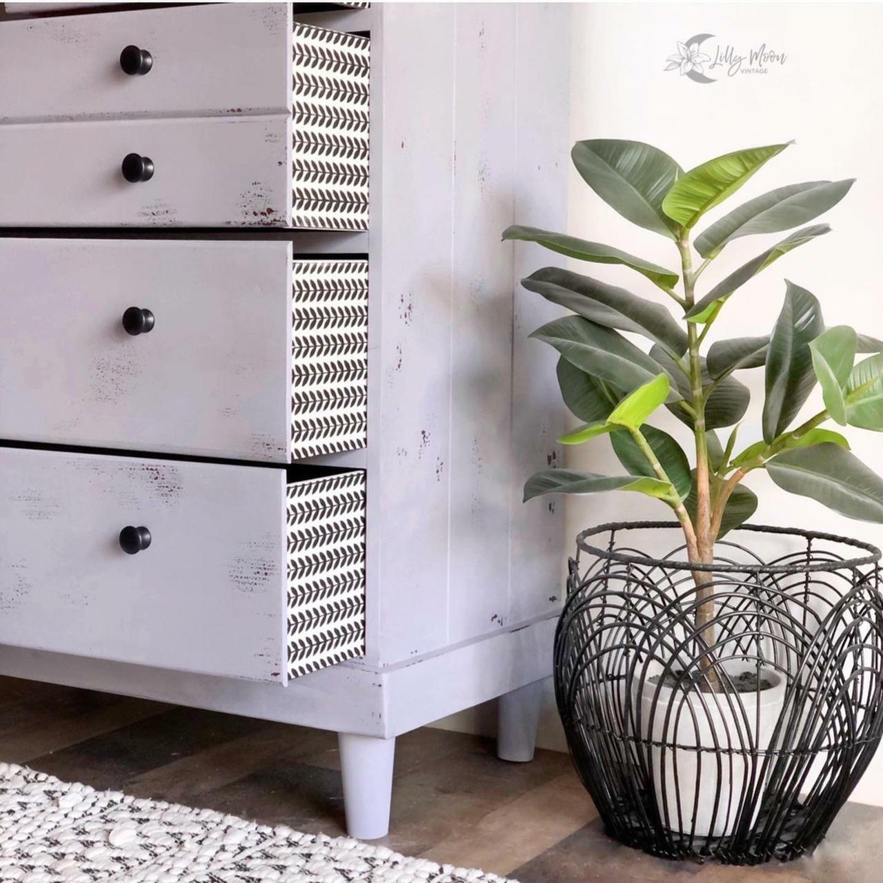 Belles and Whistles Botanical - Silkscreen Stencil provides an extra decorative touch to any project!
A dresser in a white farmhouse distressed style with opened drawers, on the side of these drawers are the Botanical - Silkscreen Stencil. Staged next to a potted plant .