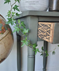 Belles and Whistles Botanical - Silkscreen Stencil provides an extra decorative touch to any project!
The side of a console table with the drawer open, on the side of these drawers is the Botanical - Silkscreen Stencil. Staged with a white bowl, greenery and a cut off Globe.