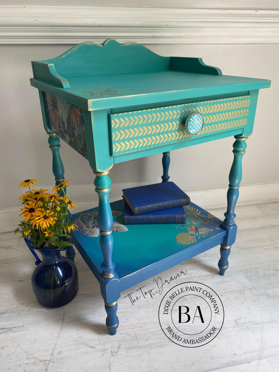Belles and Whistles Botanical - Silkscreen Stencil provides an extra decorative touch to any project!
End table with a green blended to blue paint along with Botanical - Silkscreen Stencil with gold on the front of the drawer. Staged with a blue flower vase and yellow flowers to the side of the table.