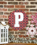 Belles and Whistles Buffalo Check - Stencil provides an extra decorative touch to any project!
A red round sign with a large white letter "P" on the background has Buffalo Check - Stencil in black. Presented on a red brick wall hanging above a row of mistletoe and a  white and red snowflake signs is seen on the right.