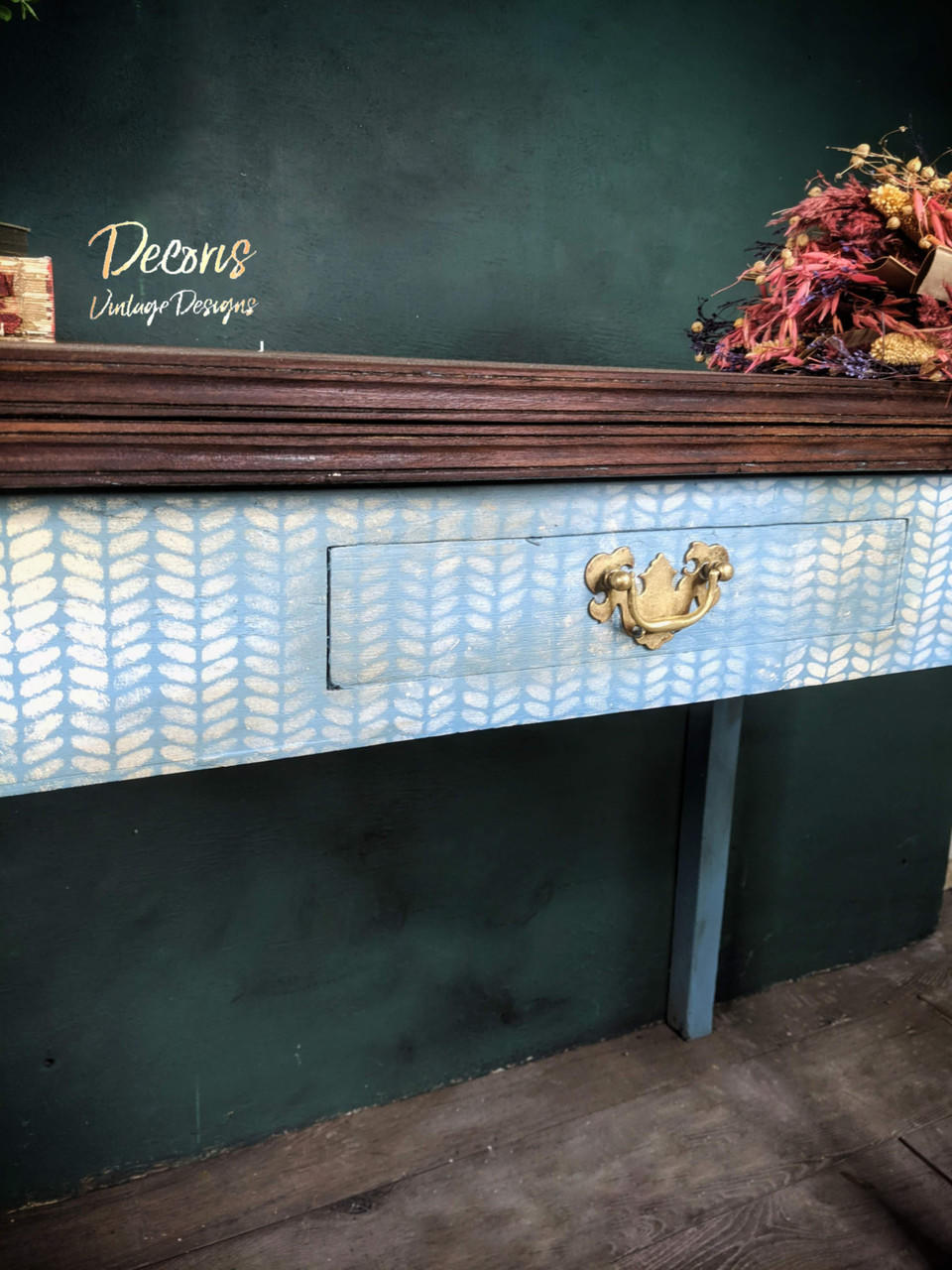 Belles and Whistles Cozy Sweater - Stencil provides an extra decorative touch to any project! A desk that has a dark wooden top and a blue base with one drawers. The desk features the cozy sweater stencil in white on the front and the drawer.