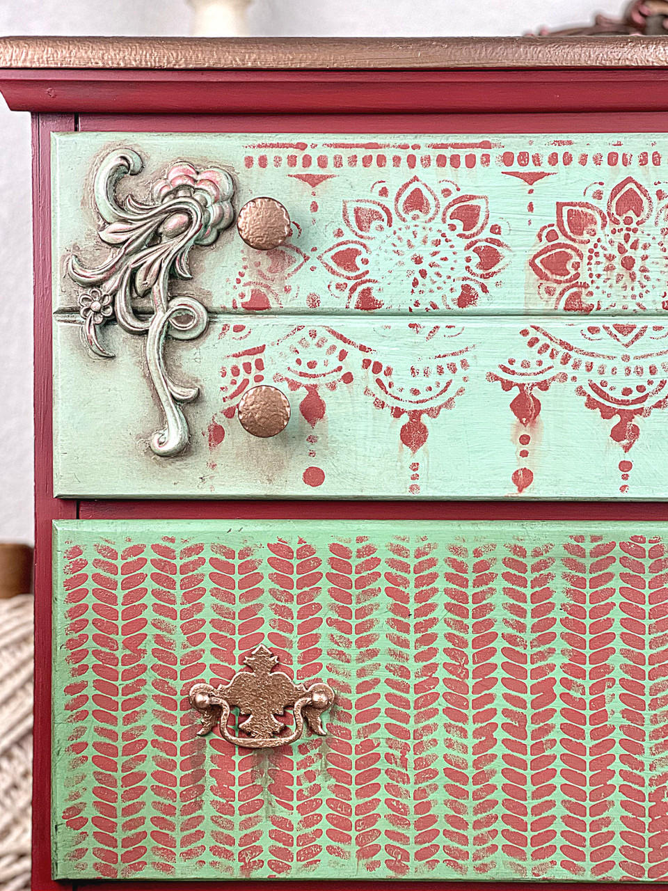 Belles and Whistles Cozy Sweater - Stencil provides an extra decorative touch to any project! A closeup image of a dresser that is painted in 2 colors with a mauve pink being on the sides and then a light blue on the drawer fronts. The first drawer has our lotus bloom stencil in the same mauve color and the second drawer features our cozy sweater stencil.
