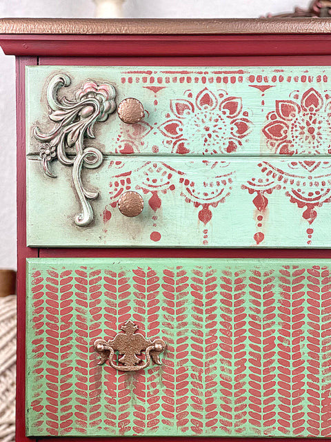 Belles and Whistles Cozy Sweater - Stencil provides an extra decorative touch to any project! A closeup image of a dresser that is painted in 2 colors with a mauve pink being on the sides and then a light blue on the drawer fronts. The first drawer has our lotus bloom stencil in the same mauve color and the second drawer features our cozy sweater stencil.
