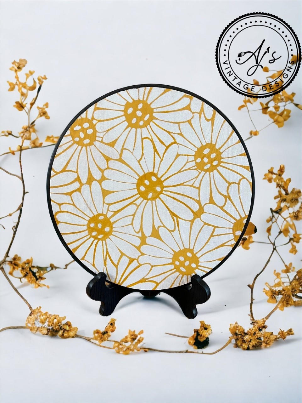 Crazy Daisy stencil applied on a plate in white and yellow with a black rim. Placed on a black plate holder. On a white background with yellow flowers strewn around.