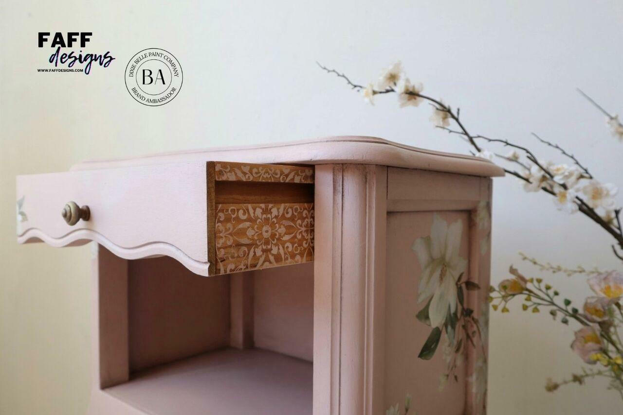 Belles and Whistles Delicate Lace - Silkscreen Stencil provides an extra decorative touch to any project!
This is a bedside table that is painted pink.  The drawer is pulled out to show the mosaic flowers stencil was drawn in white on the outside of the drawer slide. There is a big white flower decoupaged on the side of the bedside table.  There are flowers to the right of the table.