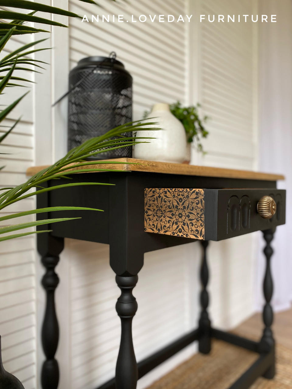 Belles and Whistles Delicate Lace - Silkscreen Stencil.
This narrow, wall table is painted black.  There is mosaic runner stencil in gold on the drawer runner.  The knob on the drawer is gold.  There is a black lantern and a plant in a white vase on top of the table .  There are fern leaves entering the picture from the left side.