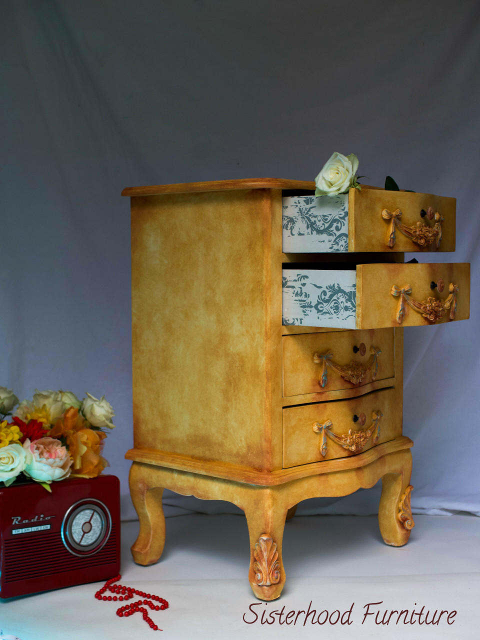 Belles and Whistles Distressed Damask - A1 Rice Decoupage Paper 
This is a beefy, mid-height dresser resting on a base with four beefy legs.  It is painted in Rebel Yellow CMP and is highly distressed.  The two top drawers are pulled out and the runners have been decoupaged.  There is lose, blue fabric draped behind the dresser.  To the left of the dresser, on the floor, is an old fashioned, red radio with flowers on top.