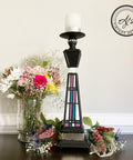 Belles and Whistles Dixie Shine 
Tall black Candle holder with Dixie shine accenting the body piece. Staged with bundles of flowers on each side of candle and a glass vase with a bundle of flowers on the left side.