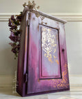 Belles and Whistles Dixie Shine 
Cabinet with a lilac and purple ombre effect with Victorian Damask - Stencil using the Dixie shine in gold on the door panel. Staged on an angle on a white wall with purple flowers and dried flower on top of piece.