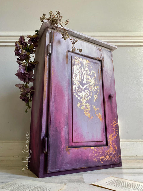 Belles and Whistles Dixie Shine 
Cabinet with a lilac and purple ombre effect with Victorian Damask - Stencil using the Dixie shine in gold on the door panel. Staged on an angle on a white wall with purple flowers and dried flower on top of piece.