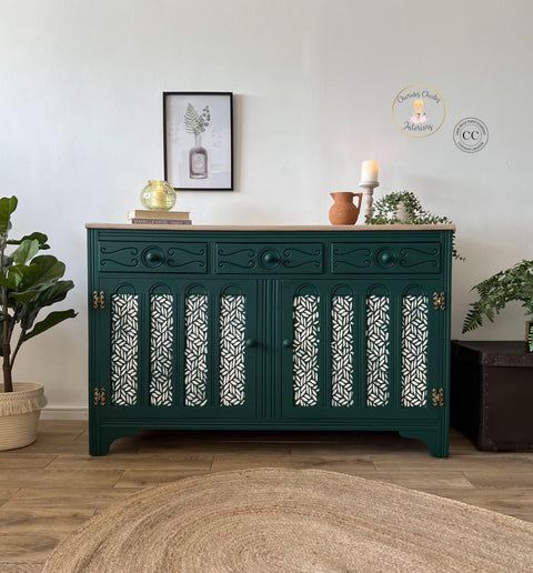 Belles and Whistles Enchanted Garden - Stencil provides an extra decorative touch to any project! Dark green painted dresser makeover with gold hinges. Enchanted Garden Stencil design applied in white on the door fronts. Staged with 2 stacked books with a short round glass vase with a candle inside. Orange terracotta pot, beige candlestick, and a beige pot containing a long vine of faux greenery.