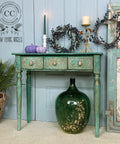 Belles and Whistles Enchanted Garden - Stencil provides an extra decorative touch to any project! Green and blue painted table with gold beetle handles on the drawers. Enchanted Garden Stencil applied on the drawer fronts. Staged with 2 purple painted pumpkins on top, a white candlestick with a long green lit candle, faux greenery & blueberry wreath, and a tall metal decorative candlestick. Large round green vase placed underneath the table with fairy lights inside.