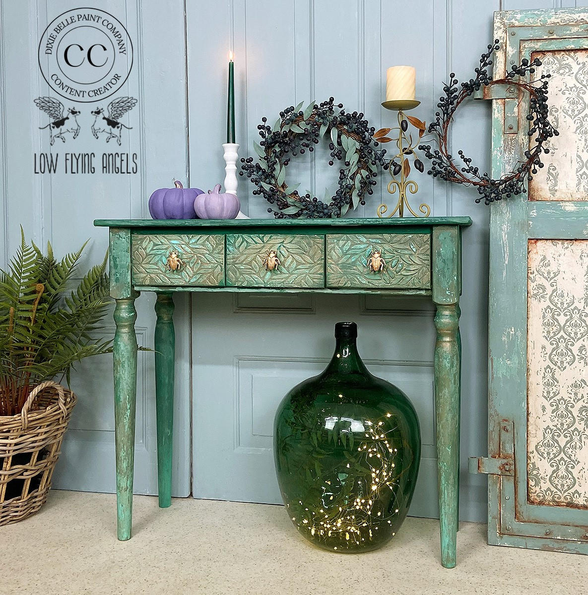 Belles and Whistles Enchanted Garden - Stencil provides an extra decorative touch to any project! Green and blue painted table with gold beetle handles on the drawers. Enchanted Garden Stencil applied on the drawer fronts. Staged with 2 purple painted pumpkins on top, a white candlestick with a long green lit candle, faux greenery & blueberry wreath, and a tall metal decorative candlestick. Large round green vase placed underneath the table with fairy lights inside.