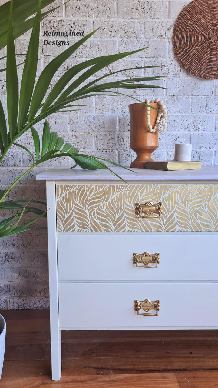 Belles and Whistles Enchanted Garden - Stencil provides an extra decorative touch to any project! White painted dresser with gold handles. Enchanted Garden Stencil applied on the top drawer front in gold. Staged with a book with gold pages on top and an orange vase with wooden beads spilling out of it.