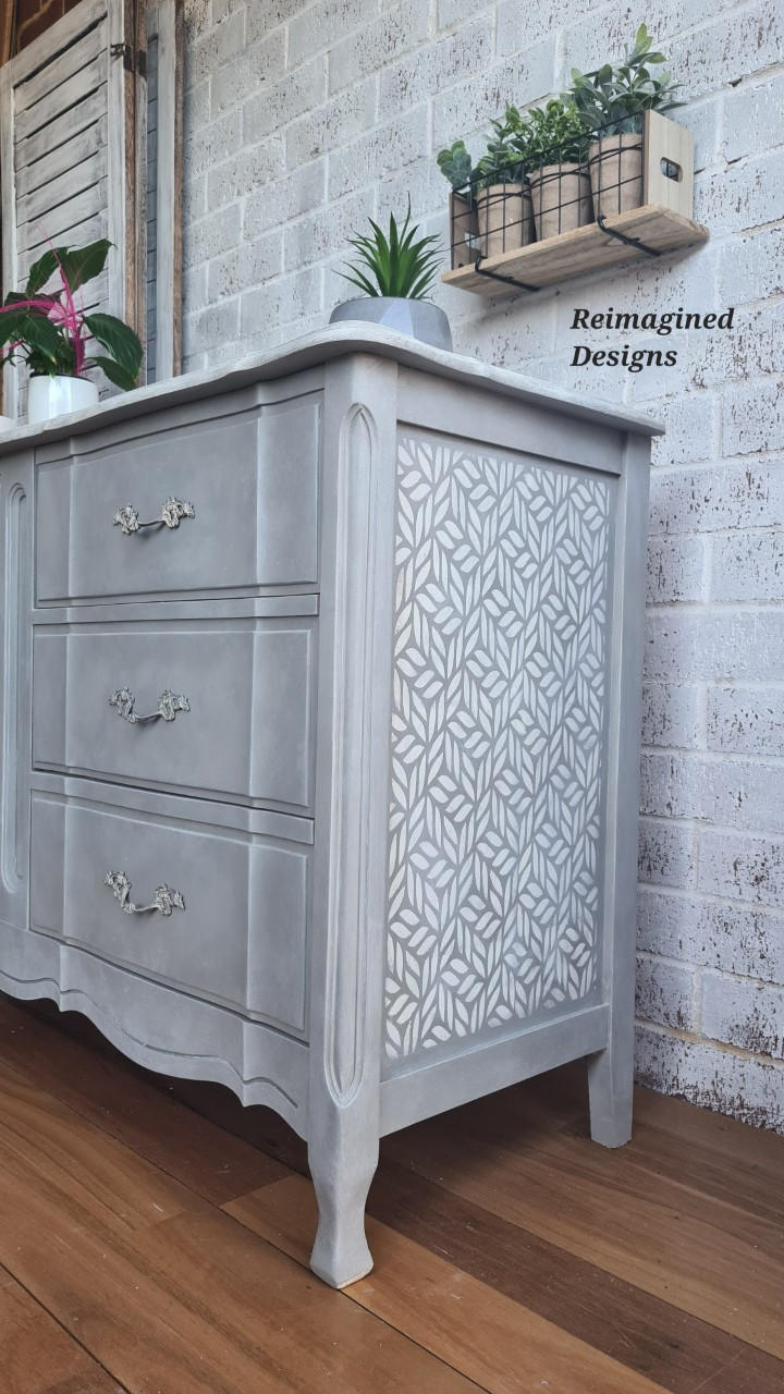 Belles and Whistles Enchanted Garden - Stencil provides an extra decorative touch to any project! Gray painted dresser makeover with one of the Enchanted Garden designs stenciled on the side in white. Staged on a wood floor with a white brick wall in the background.