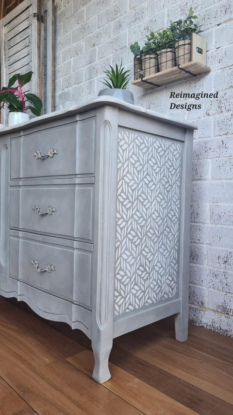 Belles and Whistles Enchanted Garden - Stencil provides an extra decorative touch to any project! Gray painted dresser makeover with one of the Enchanted Garden designs stenciled on the side in white. Staged on a wood floor with a white brick wall in the background.