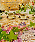 Belles and Whistles Greek Key - Stencil provides an extra decorative touch to any project! A white table with Greek Key - Stencil in gold all on top with Vintage Floral- Transfer on opposite sides of table. Staged with greenery surrounding table with two tea cups with similar patterns as Greek Key - Stencil in the center.