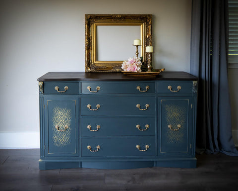 Belles and Whistles Greek Key - Stencil provides an extra decorative touch to any project! Dresser painted in a muted navy blue  with gold hardwares and Greek Key - Stencil  in gold faded a bit on the doors of the piece. Staged with a gold frame on the center of the dresser , with gold tray and two candle holder on top with some flowers.