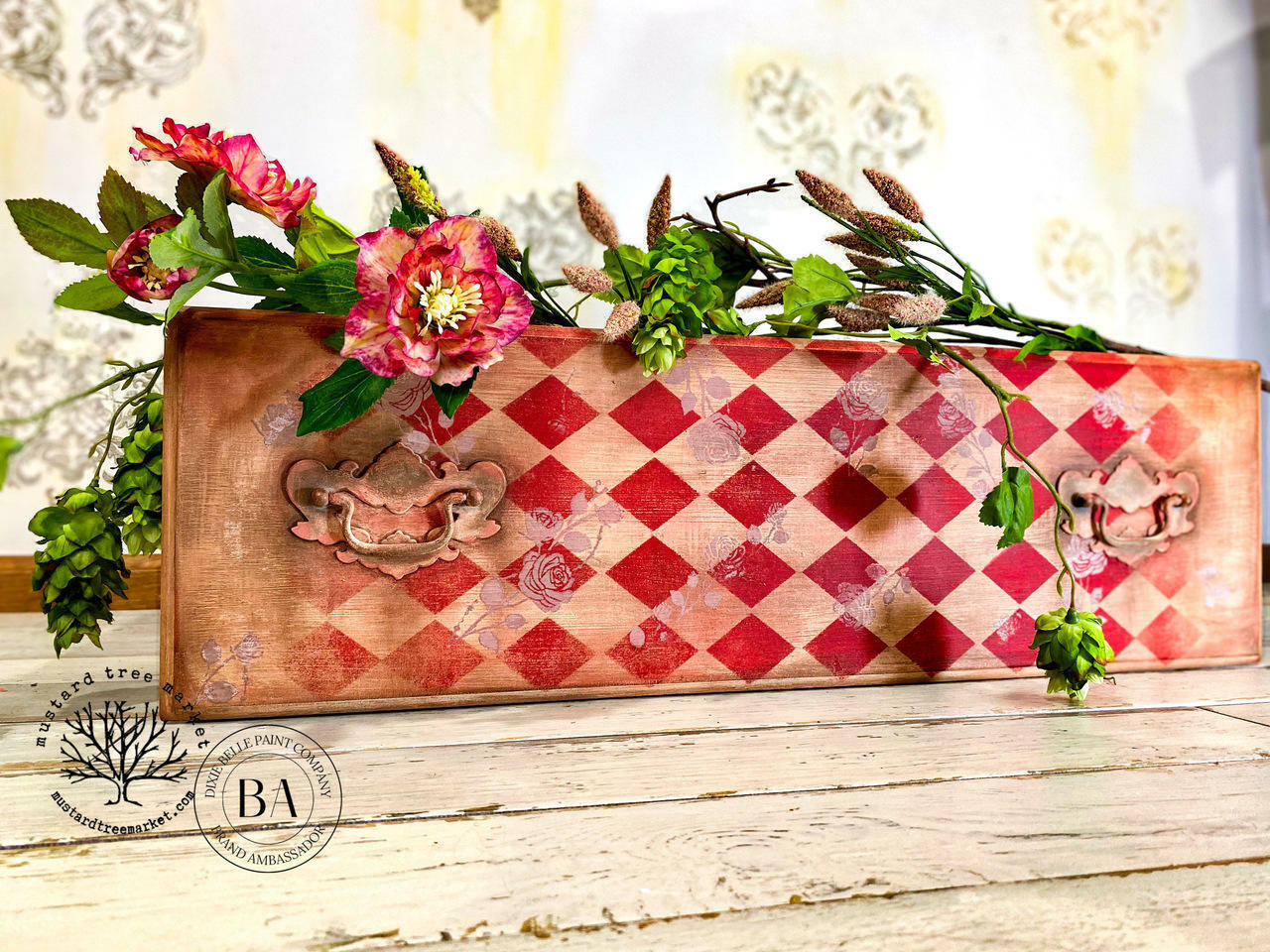 Belles and Whistles Harlequin - Stencil provides an extra decorative touch to any project!
Here's is a board with it's top facing us on a cottage, white, painted, wooden surface.  The board is stenciled with the Harlequin design in red.  Where the white would be, its left alone so you can see the board.  There are an array of flowers on top of the board.  The wall behind looks like old wallpaper that has yellowed in areas and has faded Fleur-de-leis in a brownish-gold color on the old wallpaper.