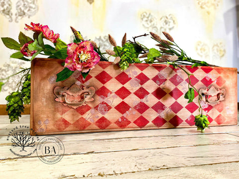 Belles and Whistles Harlequin - Stencil provides an extra decorative touch to any project!
Here's is a board with it's top facing us on a cottage, white, painted, wooden surface.  The board is stenciled with the Harlequin design in red.  Where the white would be, its left alone so you can see the board.  There are an array of flowers on top of the board.  The wall behind looks like old wallpaper that has yellowed in areas and has faded Fleur-de-leis in a brownish-gold color on the old wallpaper.