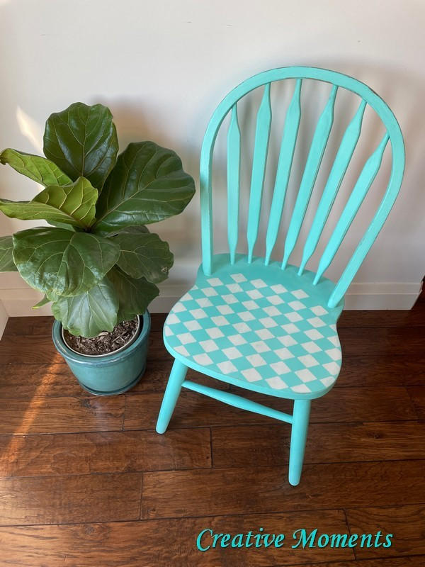 Belles and Whistles Harlequin - Stencil provides an extra decorative touch to any project!
This is a spindle chair painted turquoise.   There is a Harlequin stencil on the seat of the chair.  The colors are turquoise and white.  To the left of the chair is a potted plant.  It all rests on a slated, dark wood floor and a white wall behind.