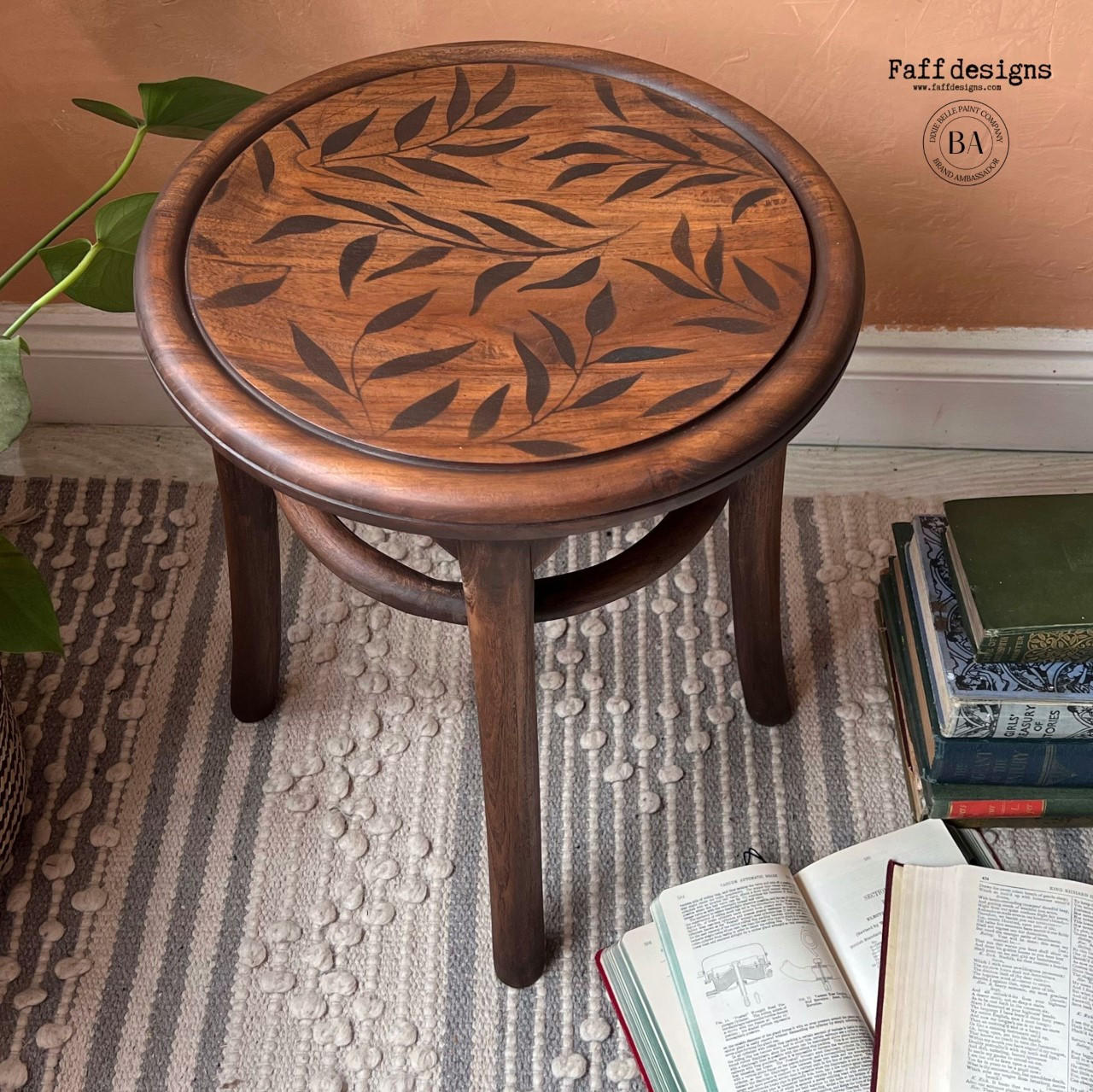 Belles and Whistles Leaf Me Alone - Stencil provides an extra decorative touch to any project! Dark wooden stool with the Leaf Me Alone stencil applied on the seat in dark brown. Staged on a white and gray rug with stacks of books around it.