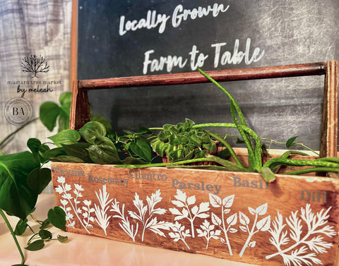 Belles and Whistles Locally Grown - Stencil provides an extra decorative touch to any project! Wooden gardening box with "Thyme, Rosemary, Cilantro, Parsley, Basil, and Dill" applied on the front top in Brown Dixie Mud. Below the names of the herbs, the corresponding stencils of the herb have been applied in White Dixie Mud. Inside the box there are multiple different plants and herbs. Behind the box there is a large blackboard with "Locally Grown" and "Farm to Table" applied in white paint.