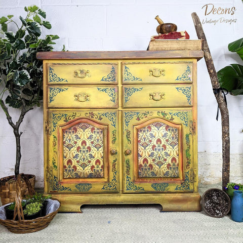 Belles and Whistles Mosaic - Silkscreen Stencil provides an extra decorative touch to any project! Yellow distressed end table with decoupage on the doors and mosaic stencil around it. Staged with plants next to it.