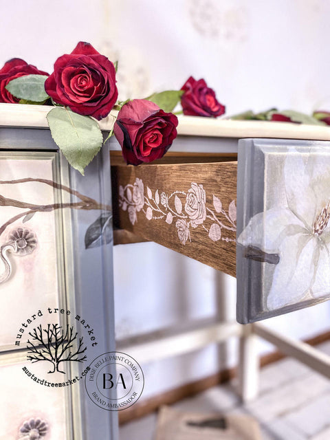 Belles and Whistles Roses - Silkscreen Stencil provides an extra decorative touch to any project! A closeup of the side of the drawers on a desk. The vertical rose design in white is shown on the the drawer side, staged with roses on top of the desk.