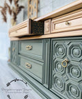Belles and Whistles Trippy Blocks - Stencil.  A green and gold chalk painted dresser with the top center drawer halfway open.  The side of the open drawer has a stencil pattern painted on it with black chalk mineral paint.  The handles and knobs are gold.  Staged on a gray floor, in front of a gray wall.  A brown plant is in the back corner.