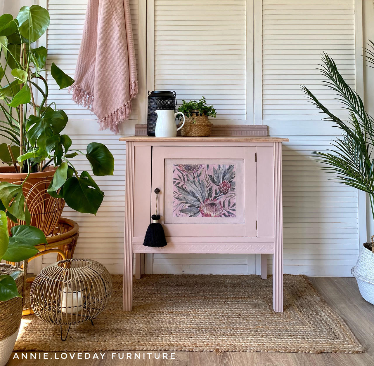 Belles and Whistles Tropical on Pink - Rice Decoupage Paper.  A pink corner table with the Tropical on Pink Rice Decoupage Paper applied to the front center of the drawer.  A black tassel hangs from the door knob.  Staged on a brown woven rug with green plants around it, and a candle inside a wooden basket.  sitting in front of bifold doors with a pink shawl hanging down.