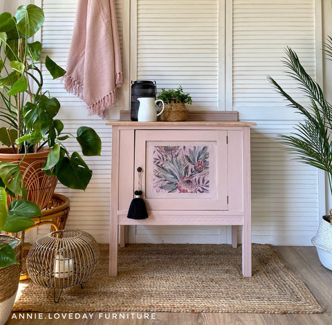 Belles and Whistles Tropical on Pink - Rice Decoupage Paper.  A pink corner table with the Tropical on Pink Rice Decoupage Paper applied to the front center of the drawer.  A black tassel hangs from the door knob.  Staged on a brown woven rug with green plants around it, and a candle inside a wooden basket.  sitting in front of bifold doors with a pink shawl hanging down.