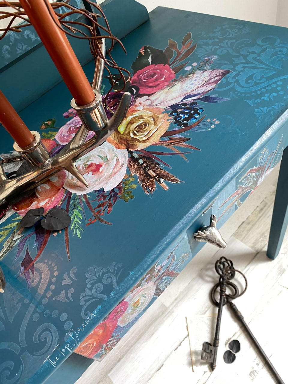 Belles and Whistles Victorian Damask - Stencil provides an extra decorative touch to any project!  The top of a blue chalk painted desk with a floral rub on transfer applied to the top and front of the desk.  The Victorian Damask Stencil is chalk painted to the top of the desk with white chalk paint.  Orange candles stand inside a gold metal candle holder on top of the desk with brown sticks weaving thru.  Brown metal keys sit on the white wooden floor.
