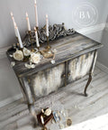 Belles and Whistles Victorian Damask - Stencil provides an extra decorative touch to any project!  A brown and white chalk painted and distressed cabinet with the Victorian Damask stencil pattern chalk painted in white to the front of the cabinet.  Staged on a light wooden floor, in front of a taupe wall with white wooden grim.  Several white candles stand in gold candlesticks on top of the cabinet, along with white roses and decorative cards and pages of books.