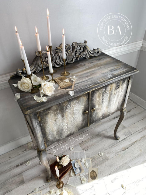 Belles and Whistles Victorian Damask - Stencil provides an extra decorative touch to any project!  A brown and white chalk painted and distressed cabinet with the Victorian Damask stencil pattern chalk painted in white to the front of the cabinet.  Staged on a light wooden floor, in front of a taupe wall with white wooden grim.  Several white candles stand in gold candlesticks on top of the cabinet, along with white roses and decorative cards and pages of books.