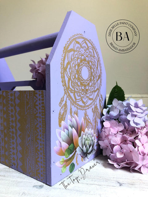 Belles and Whistles Western Boho - Silkscreen Stencil provides an extra decorative touch to any project!  A purple chalk painted magazine floor rack with the Western Boho Silkscreen Stencil applied to the front and sides with gold paint.  A muti colored succulent transfer is applied to the front.  The rack sits on a light colored wooden floor with pink and purple flowers to the right of it.