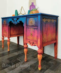 Belles and Whistles Western Boho - Silkscreen Stencil provides an extra decorative touch to any project!  Blue, pink, orange and yellow blended and layered chalk painted desk with the Western Boho Silkscreen Stencil applied to the front and sides of the desk in gold paint.  Drawer handles are gold.  The desk stands on a brown wood floor, in front of a white wall.  On top of the desk are blue, green and purple glass vases, a gold figurine, an orange flower, and a brown pot with greenery inside.