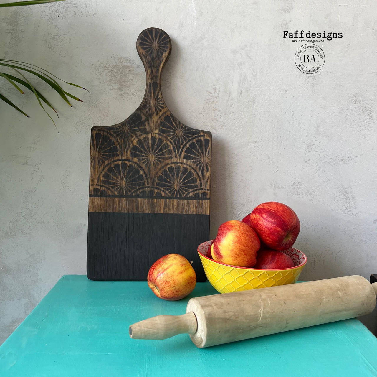 Belles and Whistles When Life Gives You Lemons - Stencil.  A wooden cutting board that is chalk painted black on the lower half and has the When Life Gives You Lemons stencil applied to the top and handle with black chalk paint.  A yellow and red bowl sits on the table holding red applies; an apple is sitting on the table in front of the cutting board; and a brown wooden rolling pin is sitting in front of the bowl.  All sits on a blue table, ion front of a gray wall.  Greenery sits at the left of the table.